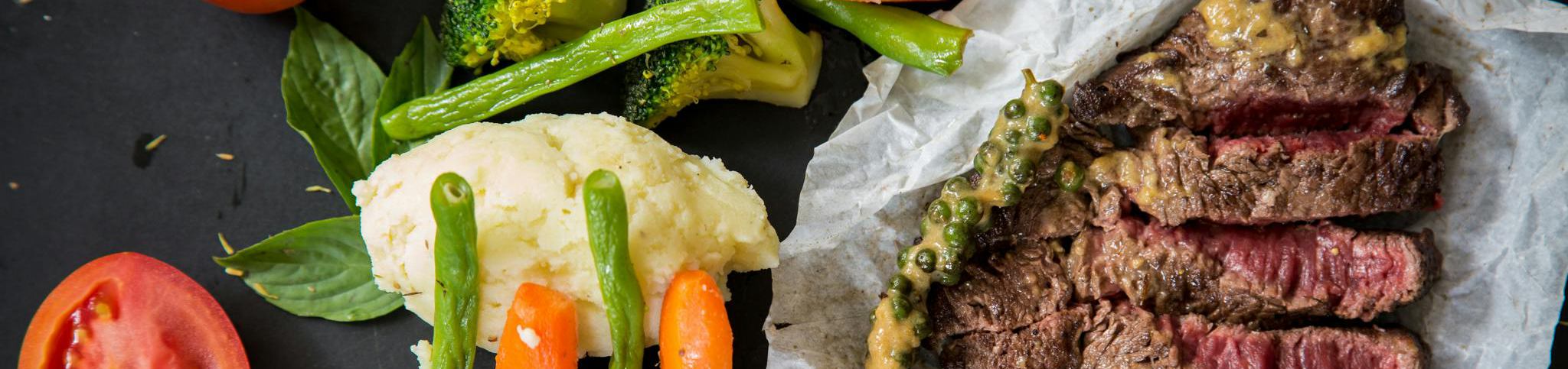 Plate with sliced medium-rare steak, mashed potatoes topped with green beans and carrots, steamed broccoli, and fresh tomato on a dark surface