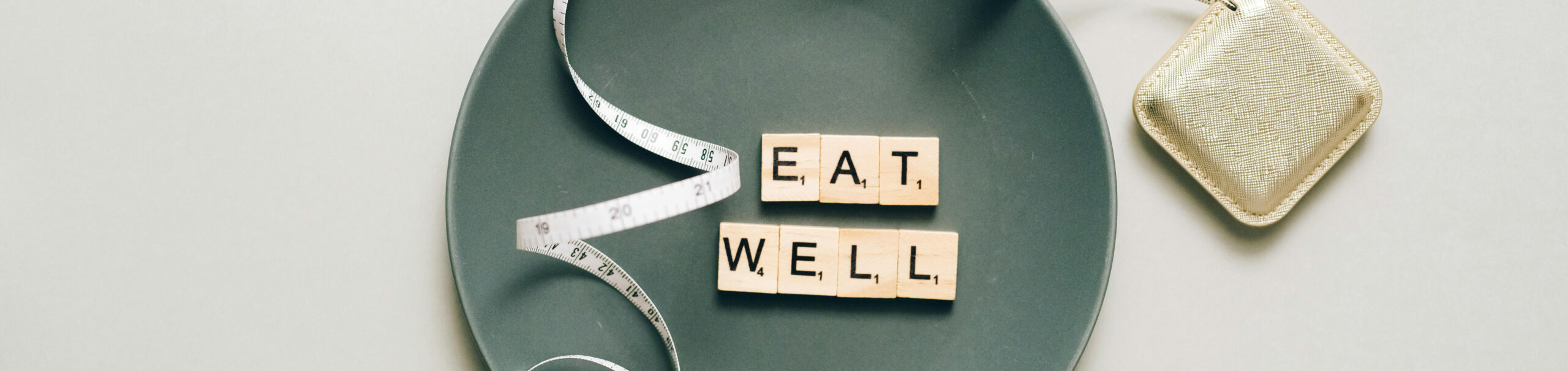 Top-down view of a gray plate with Scrabble tiles spelling “EAT WELL,” a white measuring tape draped across it, and a small gold pouch nearby.