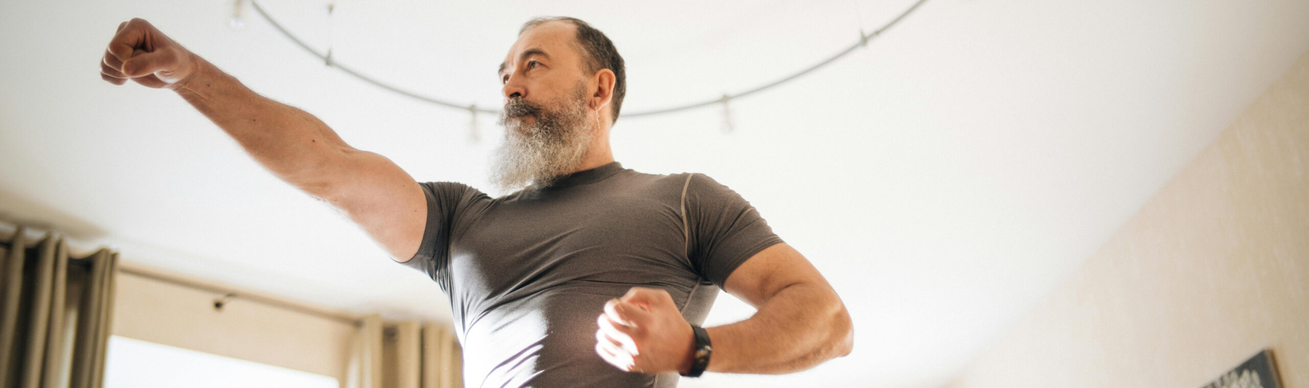 Fit older man with a gray beard striking a strong punch pose during a home workout in a bright room with curtains and natural light.