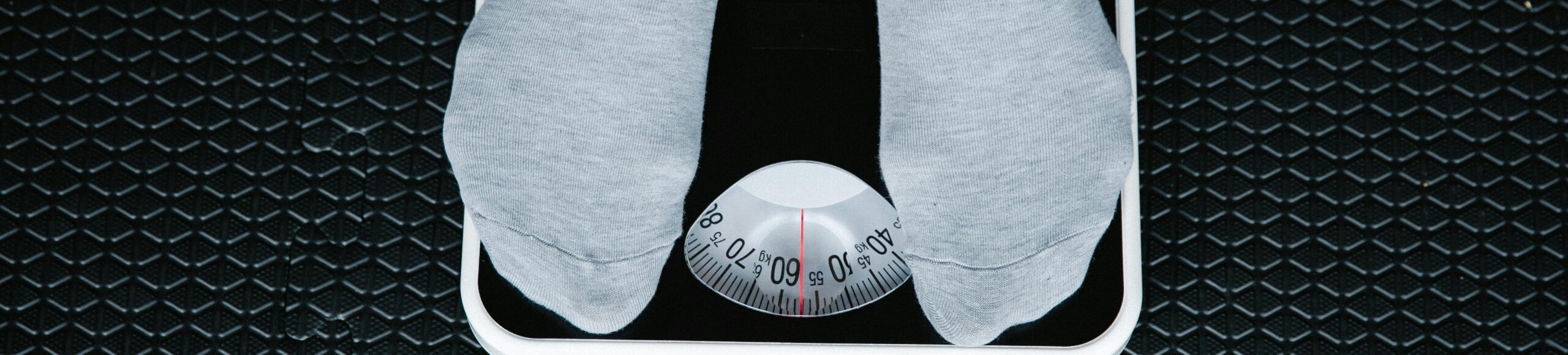 Person wearing light gray socks standing on an analog bathroom scale, showing a weight of approximately 64 kilograms, on a black textured floor.