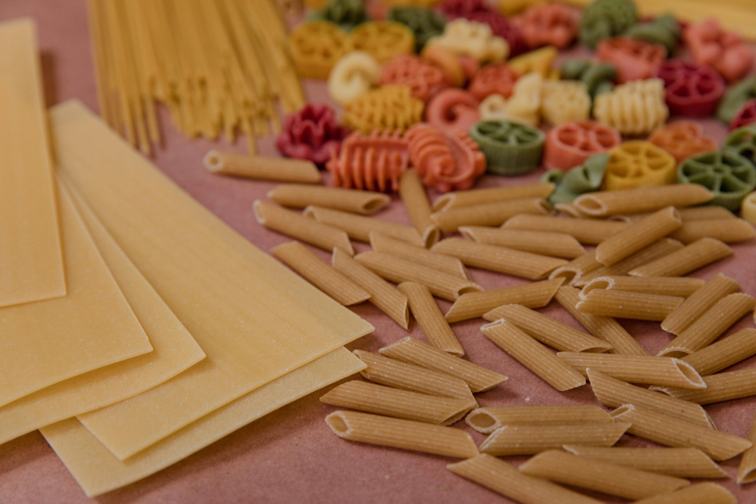 A variety of uncooked pasta types arranged on a pink surface, including penne, lasagna sheets, spaghetti, and colorful rotini-shaped pasta.