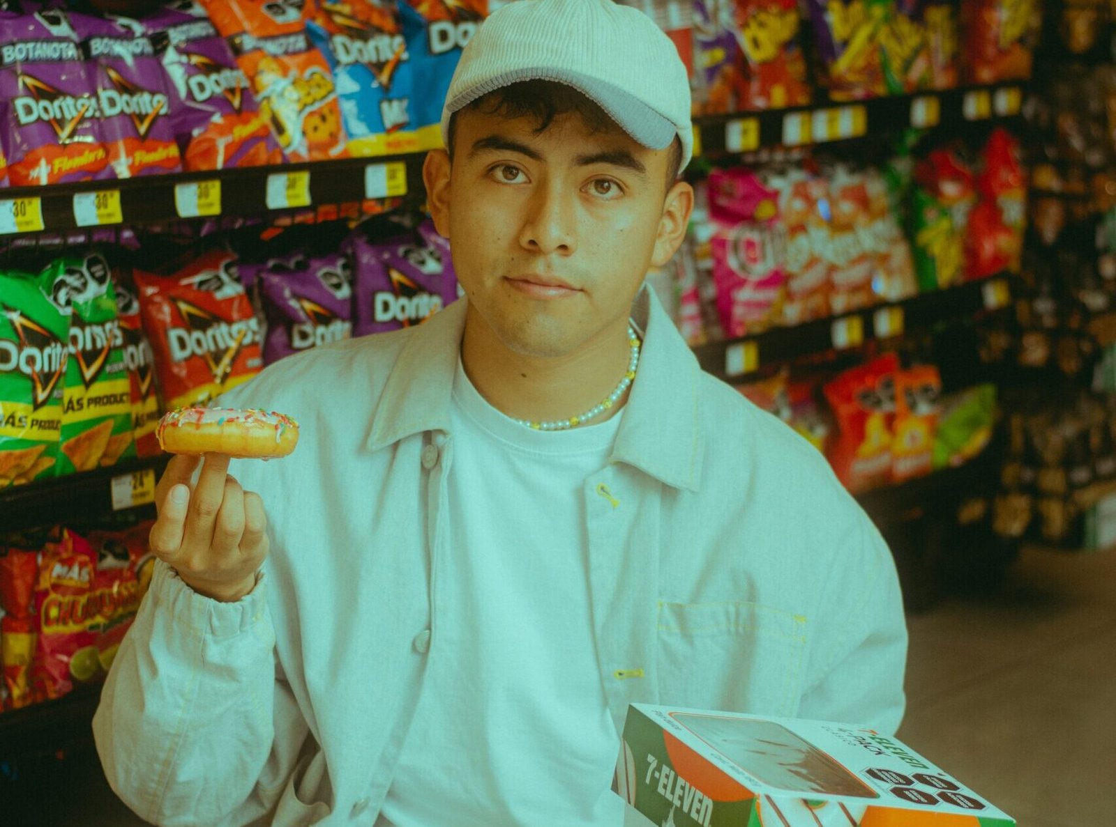 A man in a white cap and light jacket holds up a frosted snack cake in a convenience store aisle filled with colorful chips and candy.