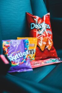 A bag of Doritos, Skittles, M&M’s, and a Pepperoni snack stick sitting on a teal car seat, representing calorie-dense snack foods or junk food.